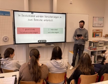 Das Bild zeigt eine Szene aus dem Schulunterricht mit Lehrer, Schüler und digitaler Tafel.